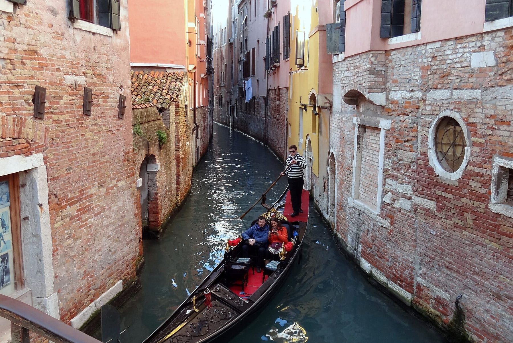 Couple seated low in a black gondola on a narrow Venetian canal, gondolier rowing behind them
