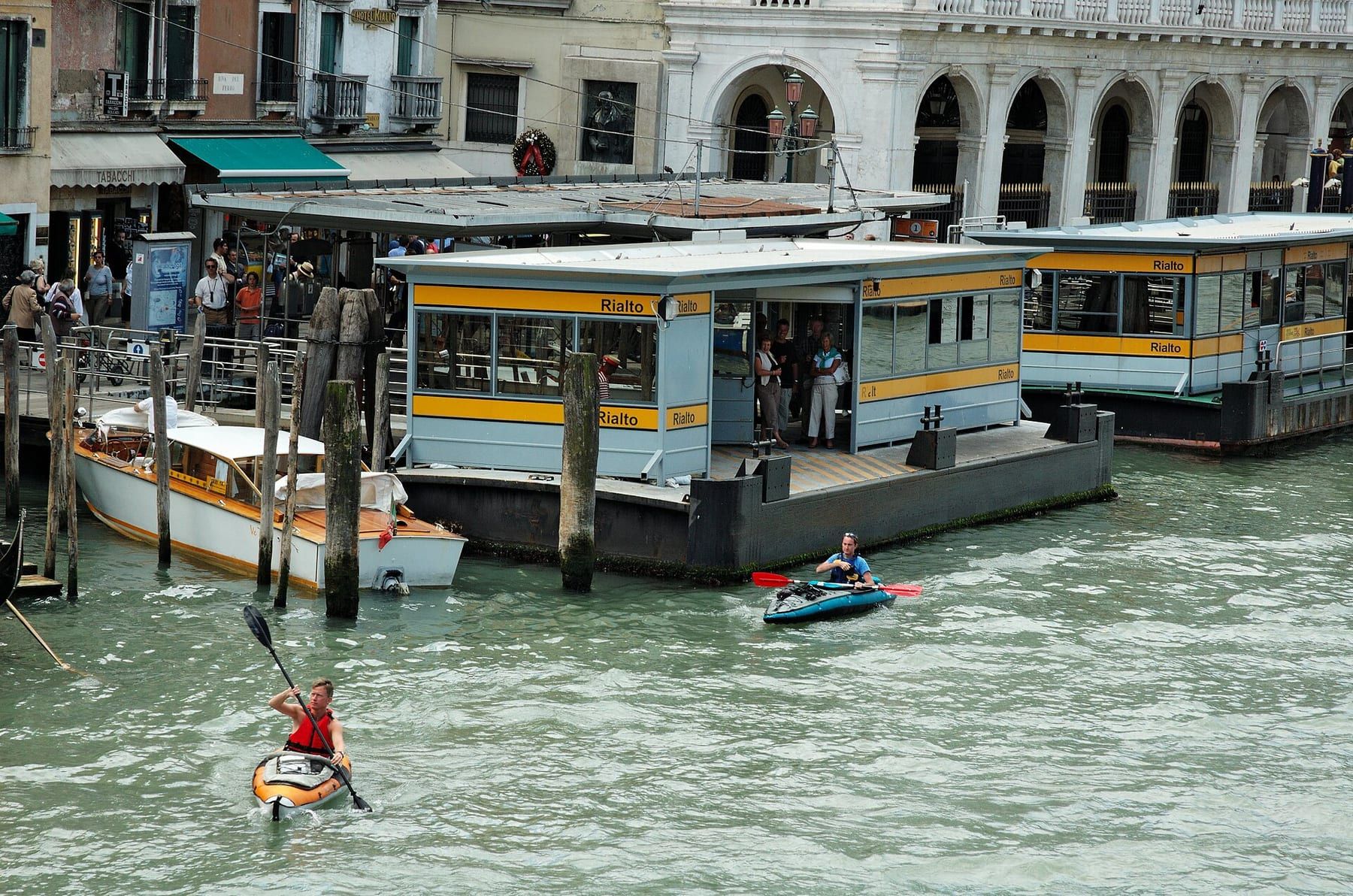 Kayakers paddling in single file along a narrow Venetian side canal beneath a low stone bridge